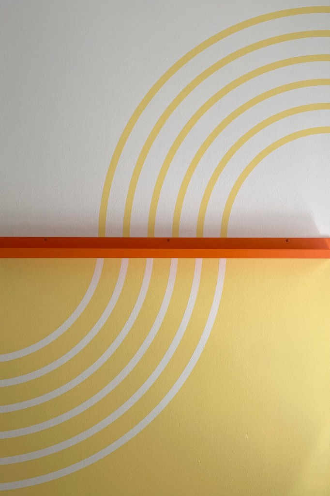 Wandmalerei in sanften Gelb- und Grautönen mit weißen Linien, dazwischen eine orange Bilderleiste.