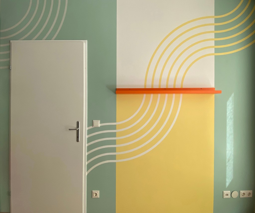 Wandgestaltung in sanften Gelbtönen, Grün und Grau mit weißen und gelben Linien und einem leuchtenden orangefarbenen Regal.