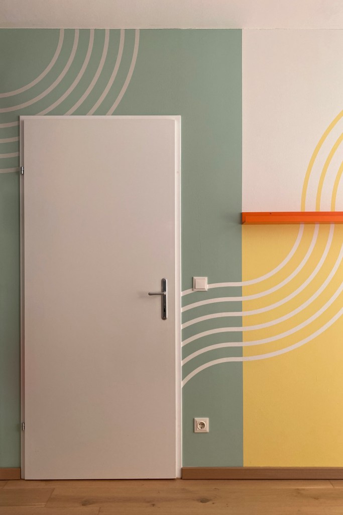 Ein Raum mit einer weißen Tür, dekorierten Wänden in sanften Grüntönen und einem gelben Abschnitt, auf dem geschwungene Linien in Weiß zu sehen sind. Eine schmale orangefarbene Regalleiste ist ebenfalls an der Wand angebracht.